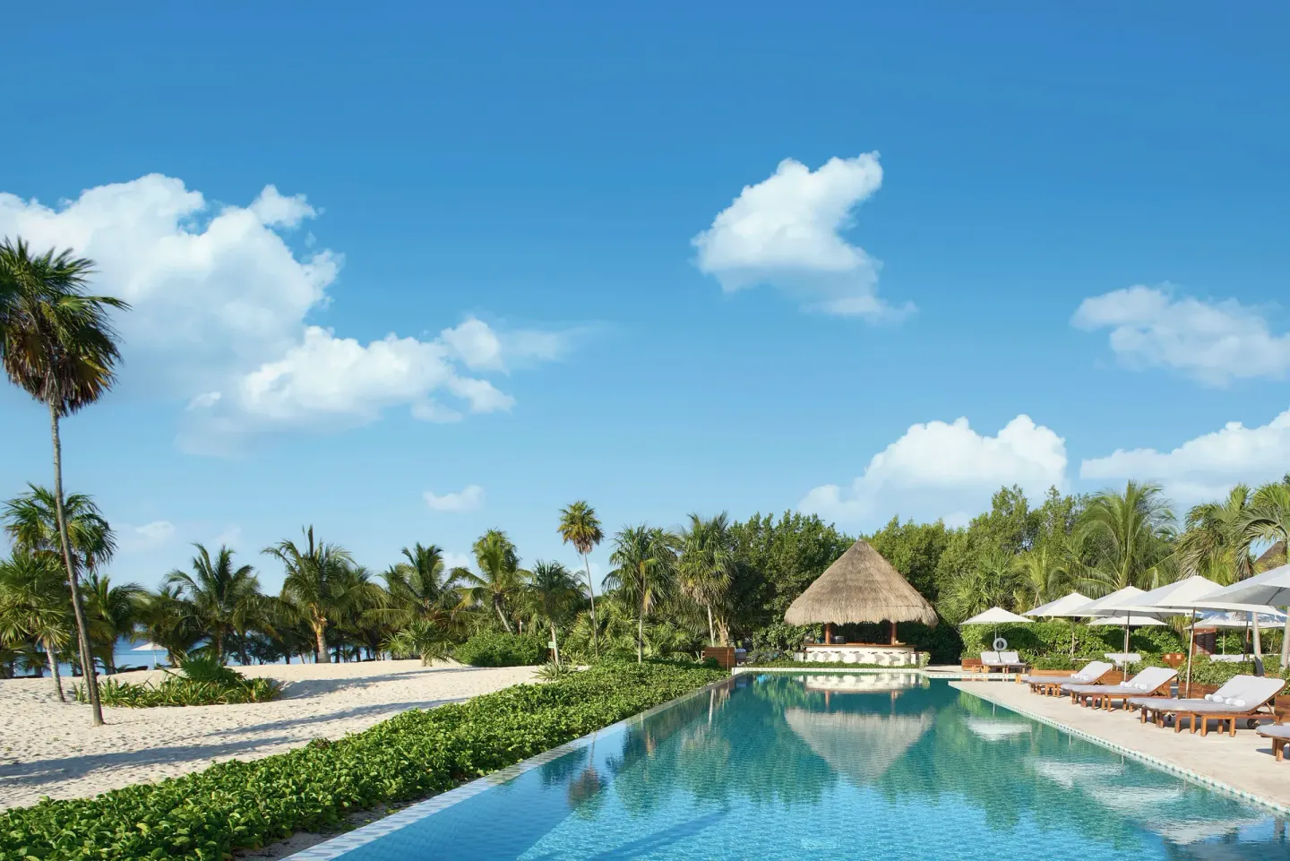 Long pool beside sand, palms, umbrellas, and a palapa at Chablé Maroma.
