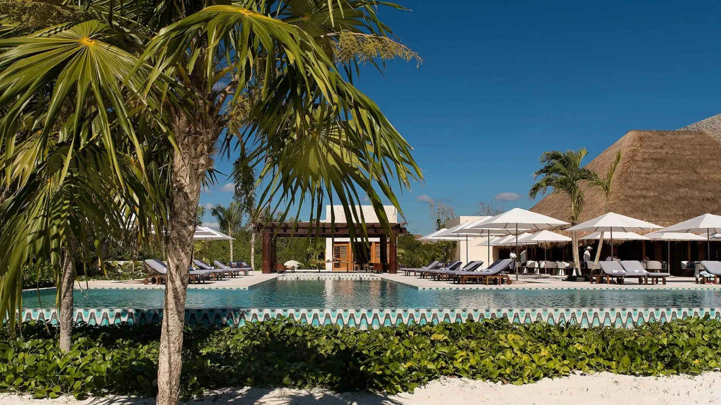Resort pool with palms, umbrellas, and a thatched-roof building at Chablé Maroma.