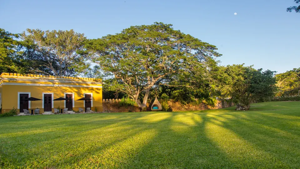 Wide lawn with a yellow building and shaded seating under a tree at Chablé Yucatán
