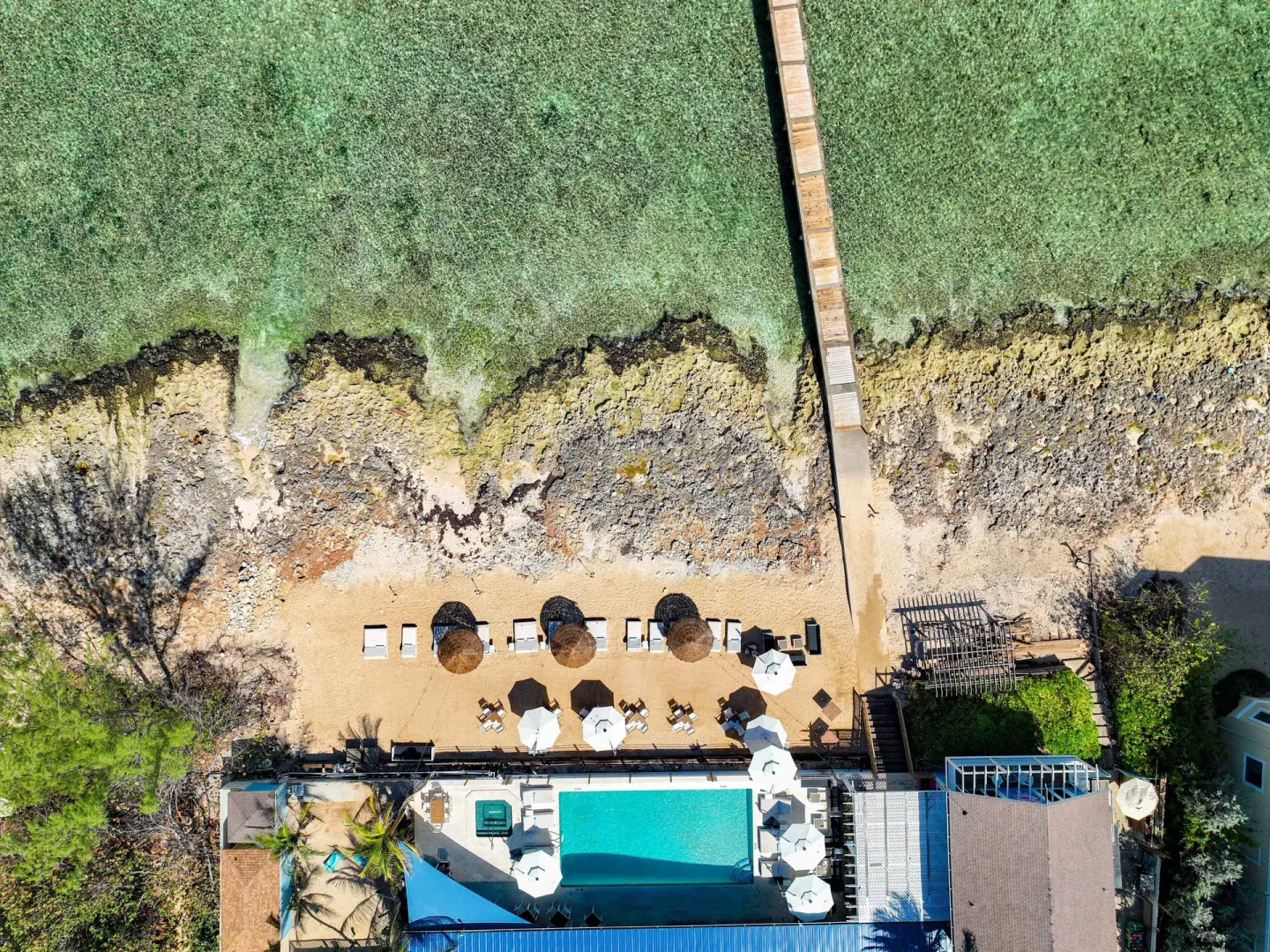 Top-down aerial view of the pier, rocky shore, pool, and beach seating at Cobalt Coast Resort.
