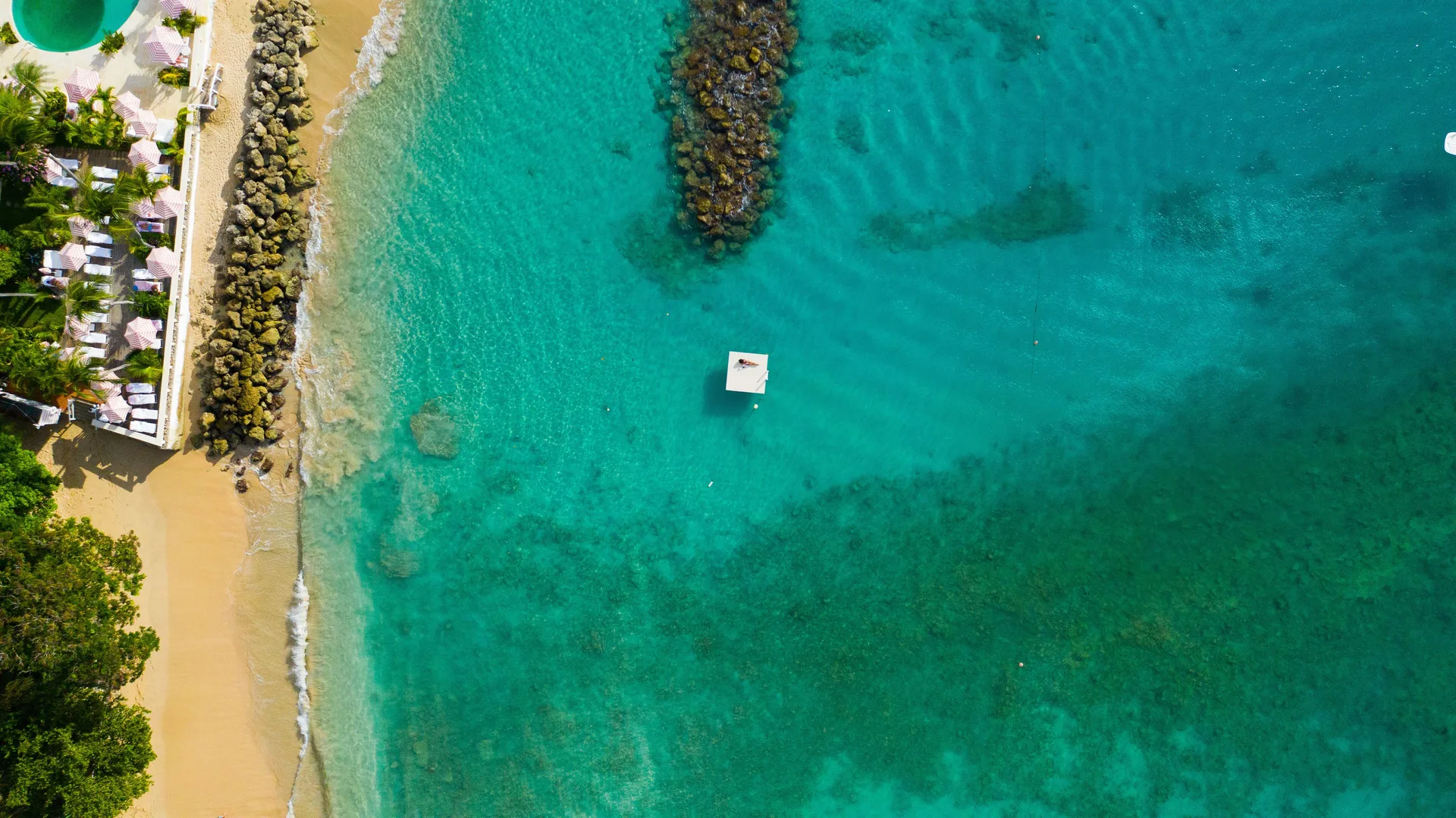 An aerial view shows the sandy beach, rock groynes, and turquoise water at Cobblers Cove.