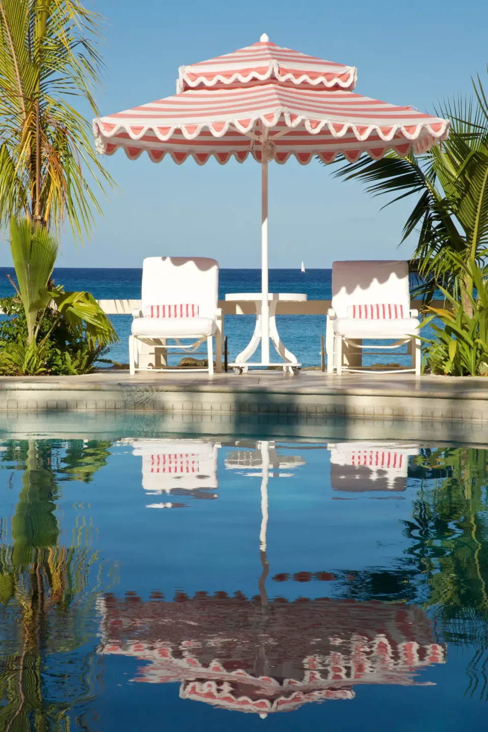 Poolside loungers and pink striped umbrella reflected in the water at Cobblers Cove.