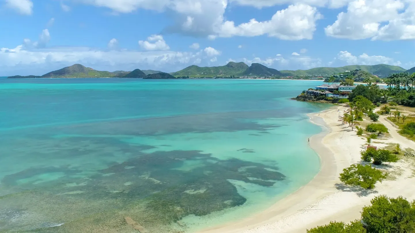 Aerial view of a curving beach, turquoise bay, and distant hills at Cocobay Resort.