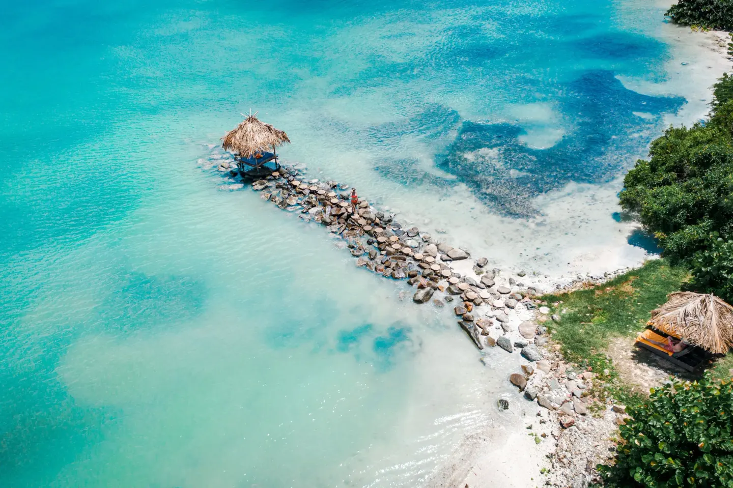 Aerial view of a rocky jetty and thatched cabanas over turquoise water at Cocobay Resort.