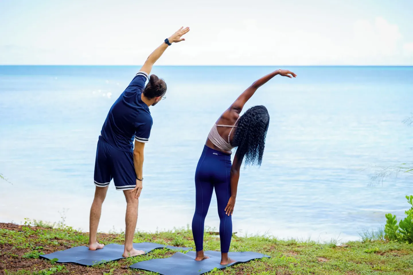Two guests side stretching on yoga mats beside the sea at Cocobay Resort.