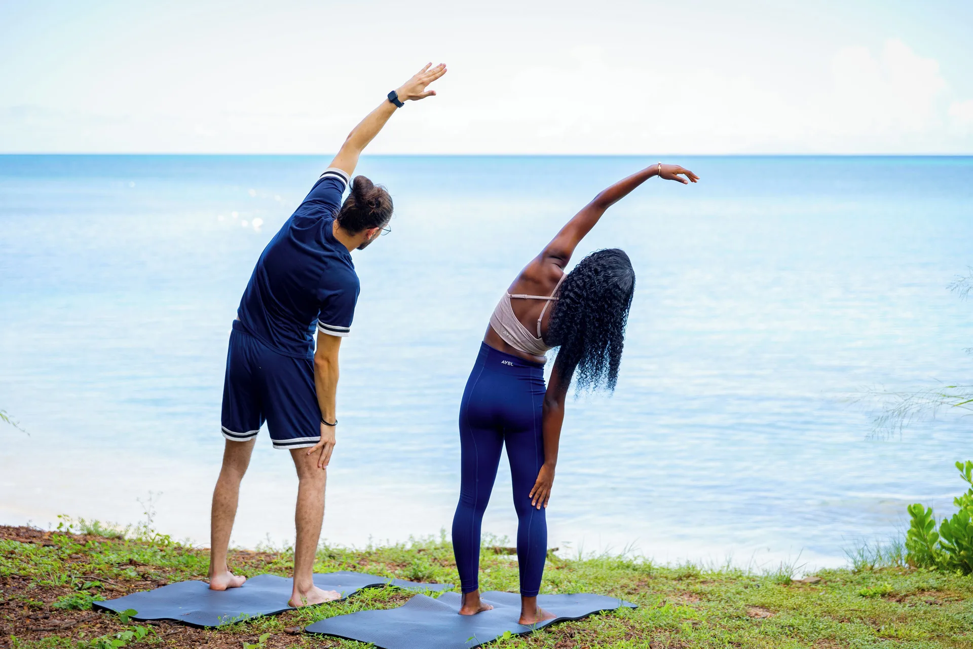 Two guests side stretching on yoga mats beside the sea at Cocobay Resort.