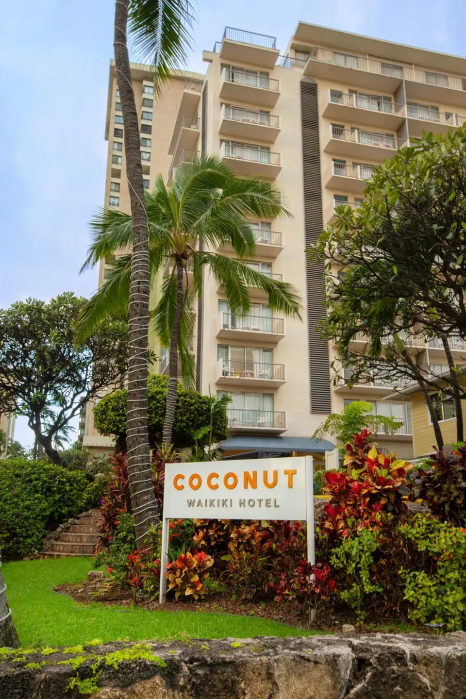 Palm-framed hotel exterior and landscaped sign at Coconut Waikiki Hotel.