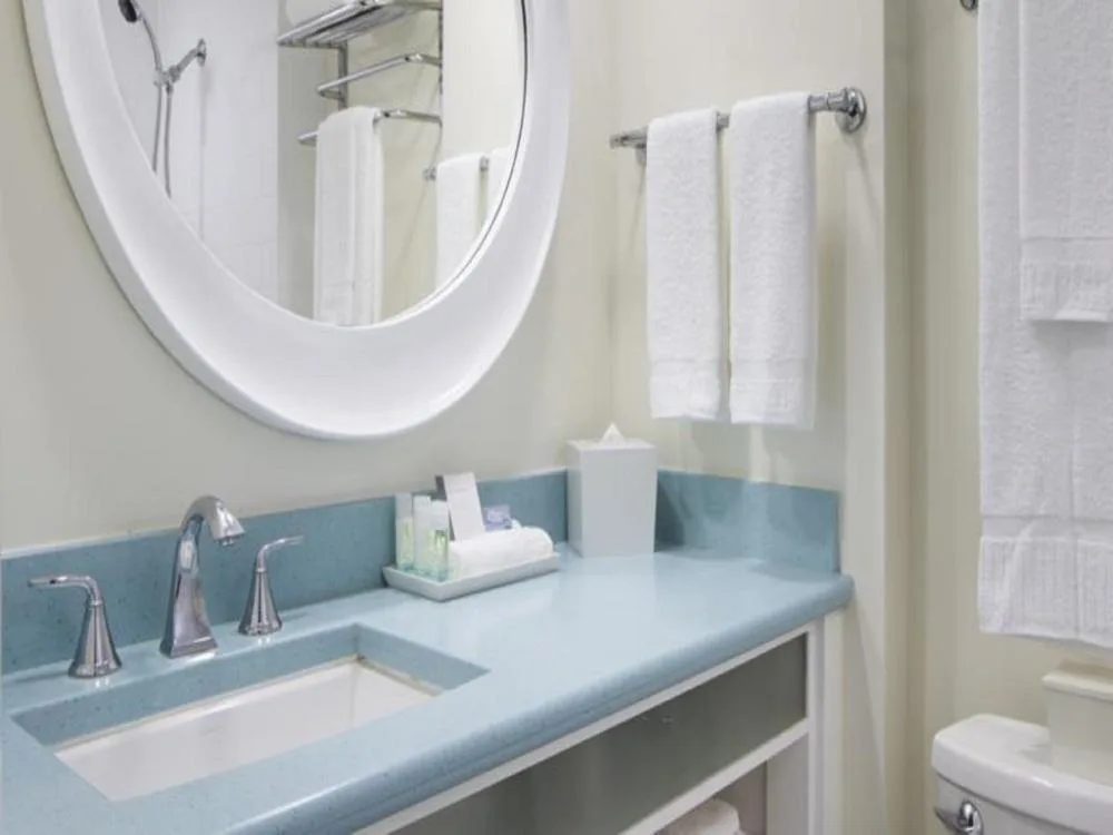 Mountain View Room bathroom at Coconut Waikiki Hotel with a blue vanity, round mirror, and white towels.