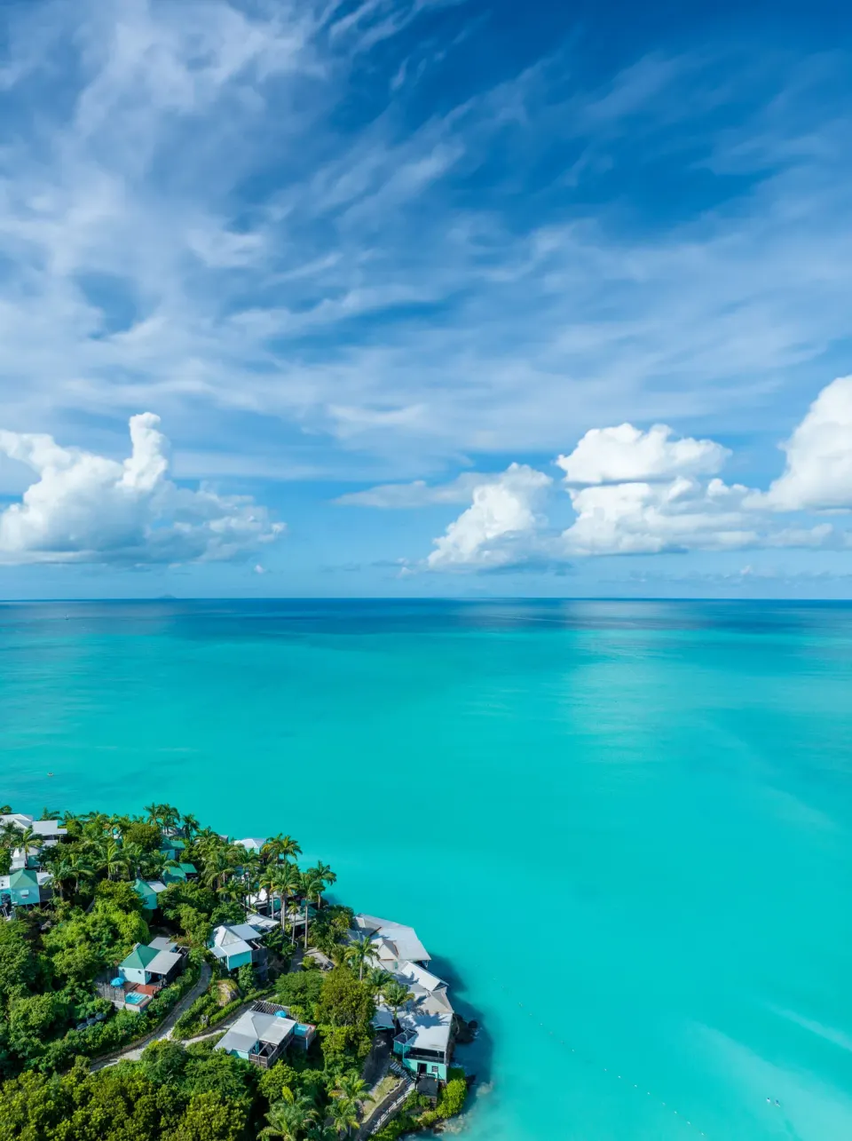Aerial view of COCOS Hotel cottages at the edge of a wide turquoise sea under cloud-filled sky.