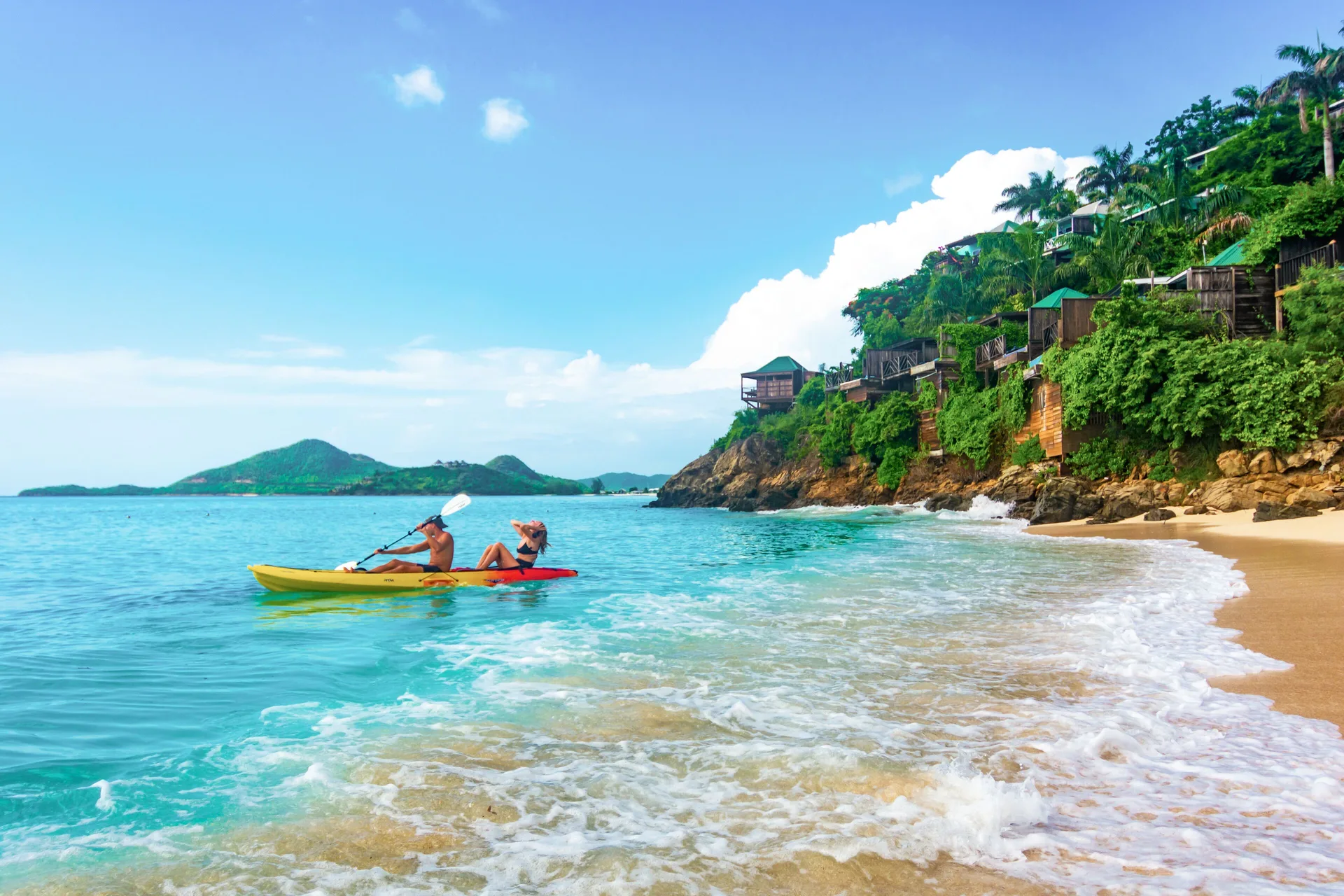 Two guests kayak beside the beach and hillside cottages at COCOS Hotel.