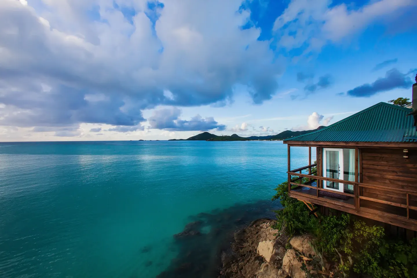A wooden cottage sits above turquoise water on the rocky coast at COCOS Hotel.