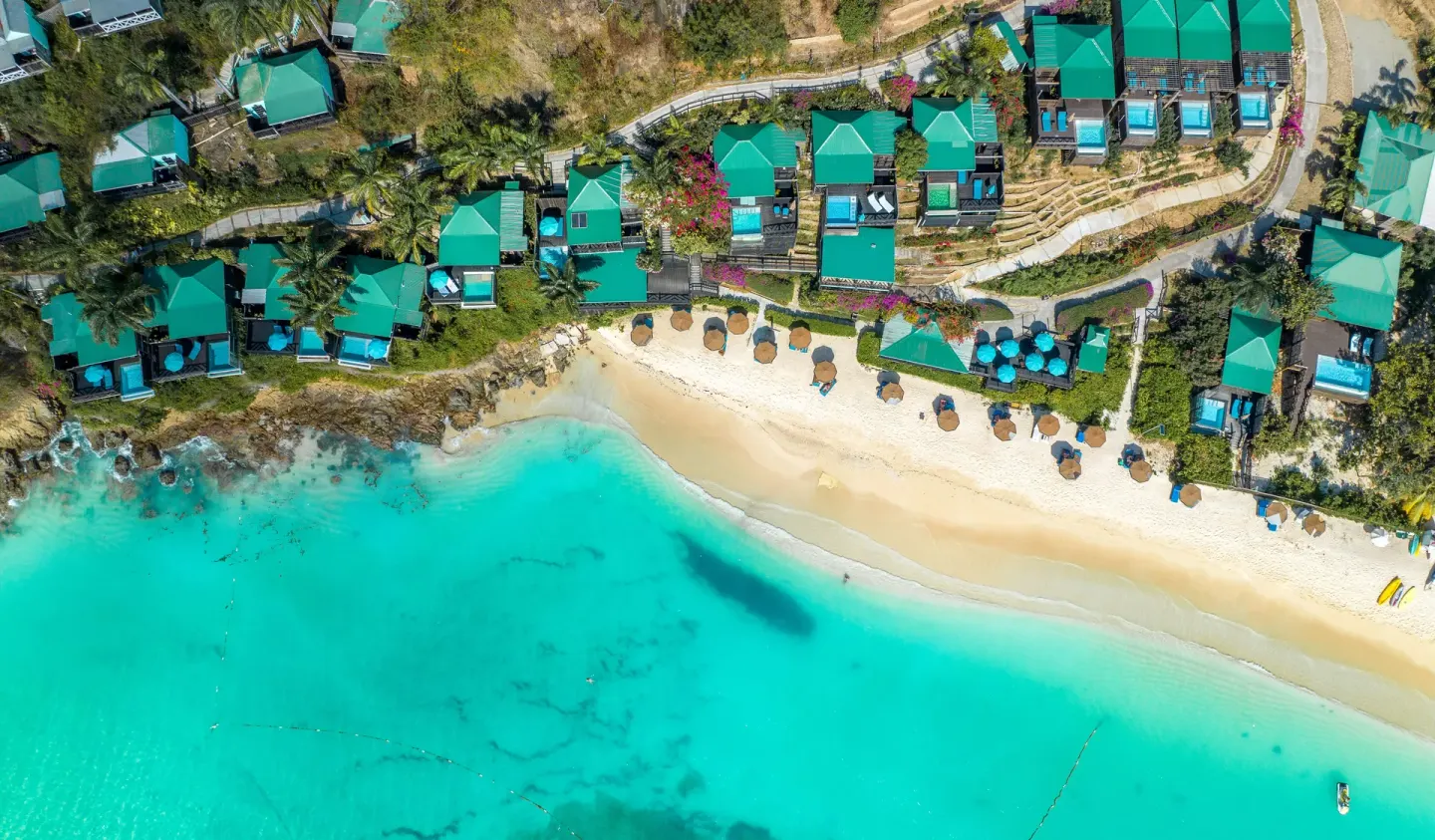 Overhead view of beach umbrellas, cottages, and clear water at COCOS Hotel.