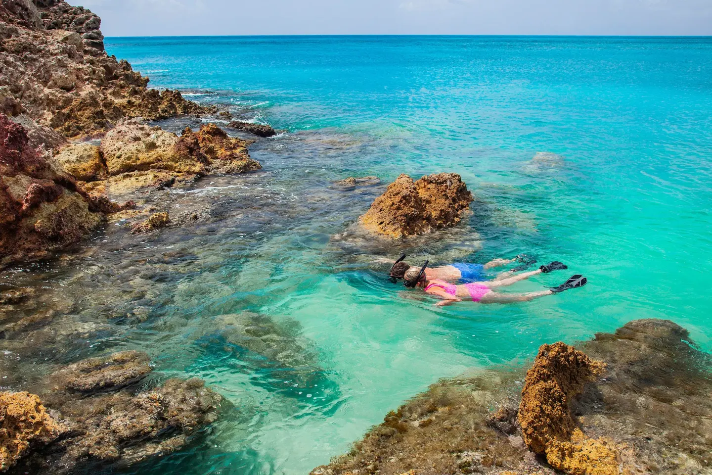 Snorkeler swimming beside rocky turquoise shoreline at COCOS Hotel.
