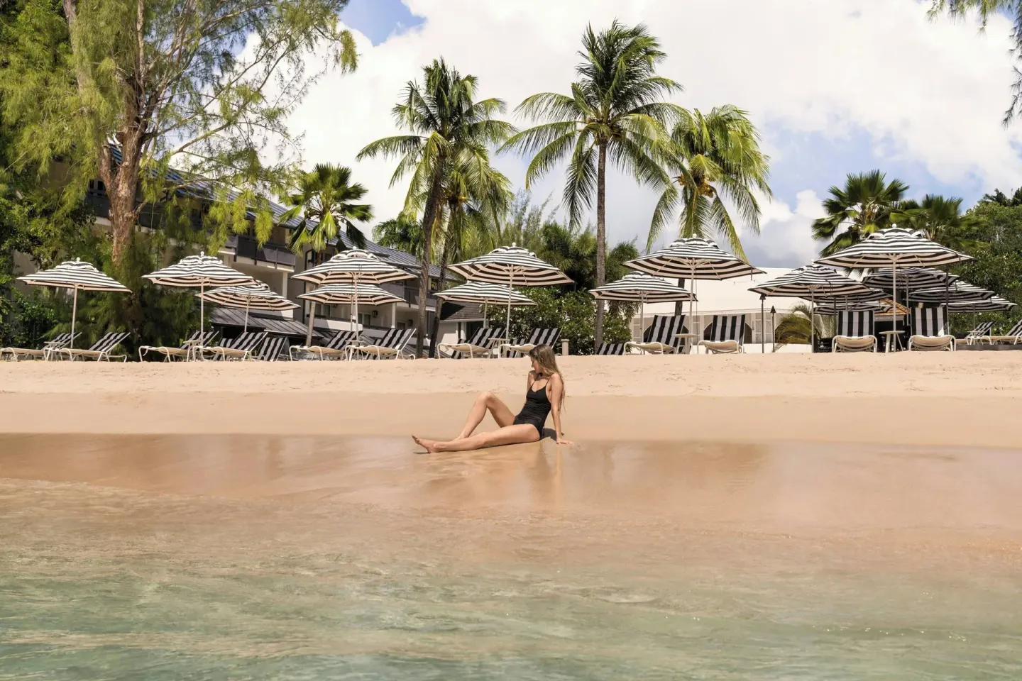 A guest sits at the shoreline below beach umbrellas and palms at Colony Club, Barbados.