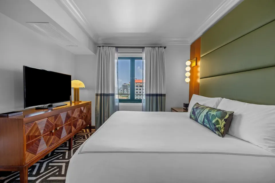 King bed, dresser, TV, and city window in the Commodore City View Suite at Condado Vanderbilt Hotel.