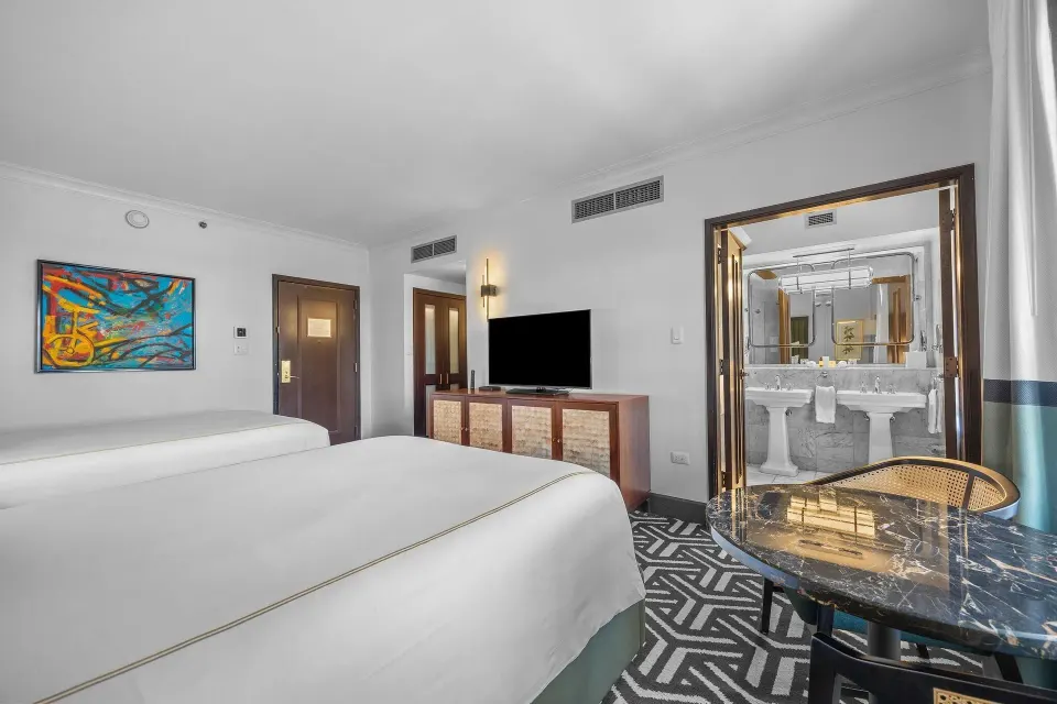 Heritage Queen Room with two queen beds, TV console, table, and mirrored bathroom view at Condado Vanderbilt Hotel.