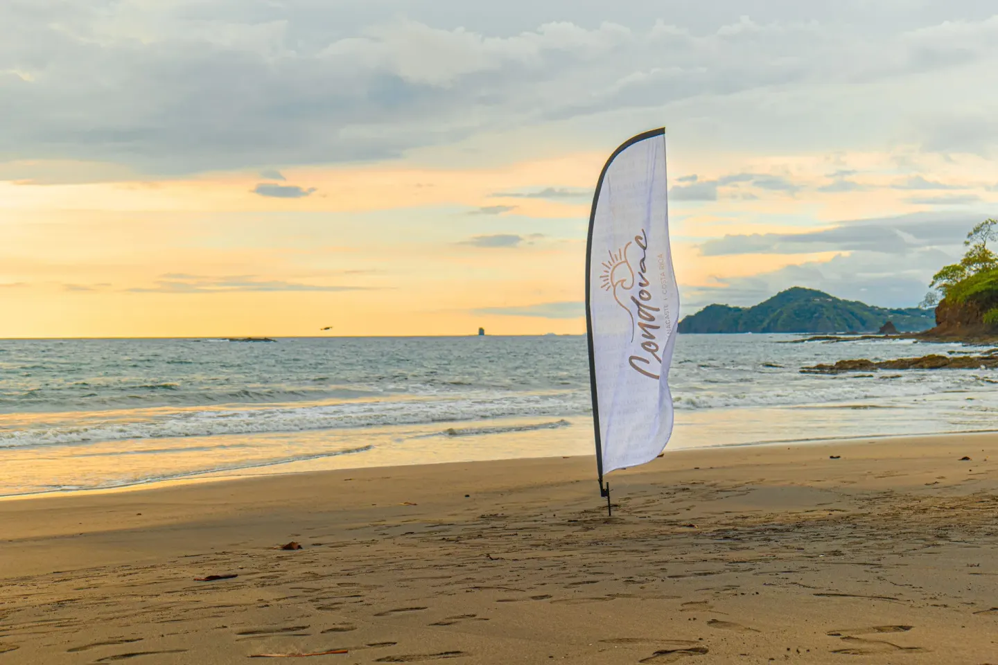 A Condovac la Costa beach flag stands in the sand near the ocean at sunset.