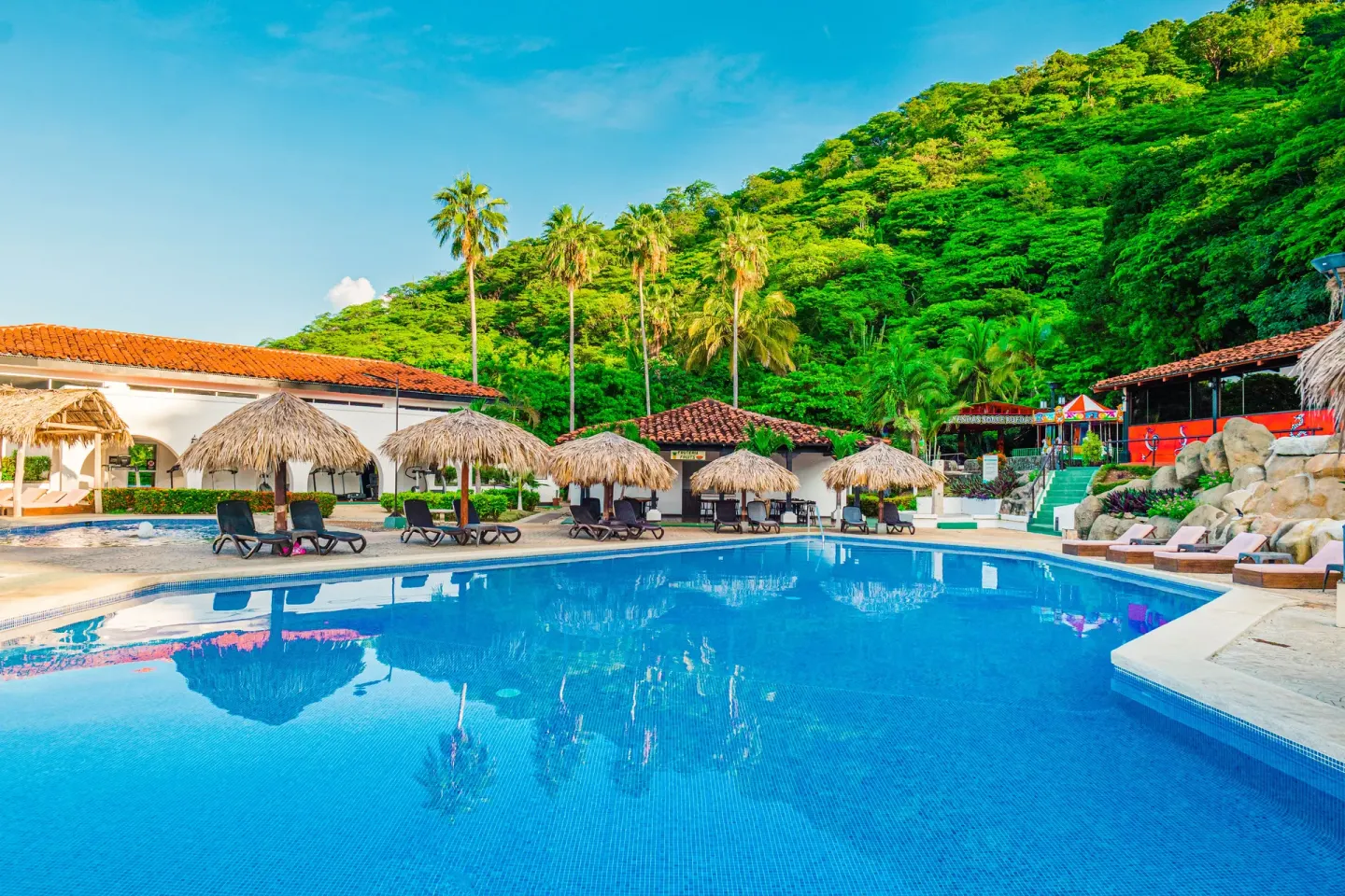Large resort pool with palapa umbrellas and hillside greenery at Condovac la Costa.