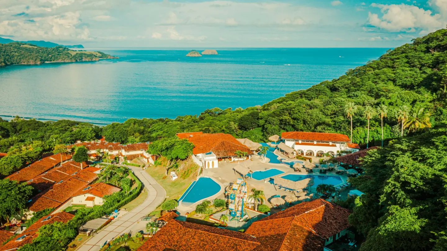 Aerial view of pools, red-roofed buildings, forest, and bay at Condovac la Costa.