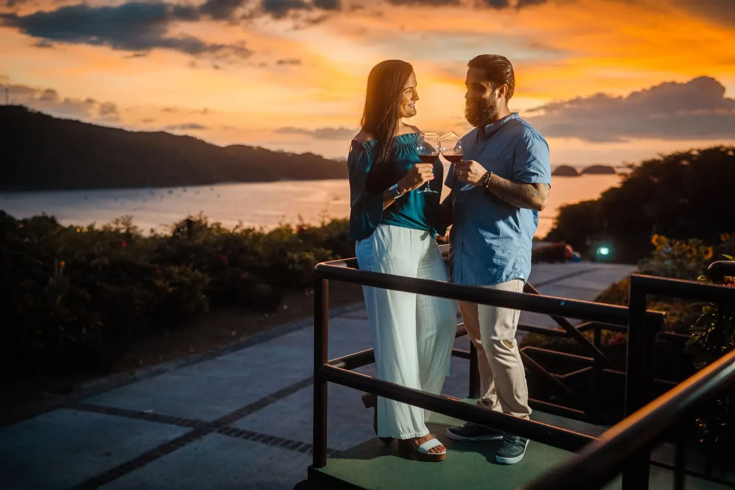 A couple holds drinks on a terrace at sunset overlooking the bay at Condovac la Costa.