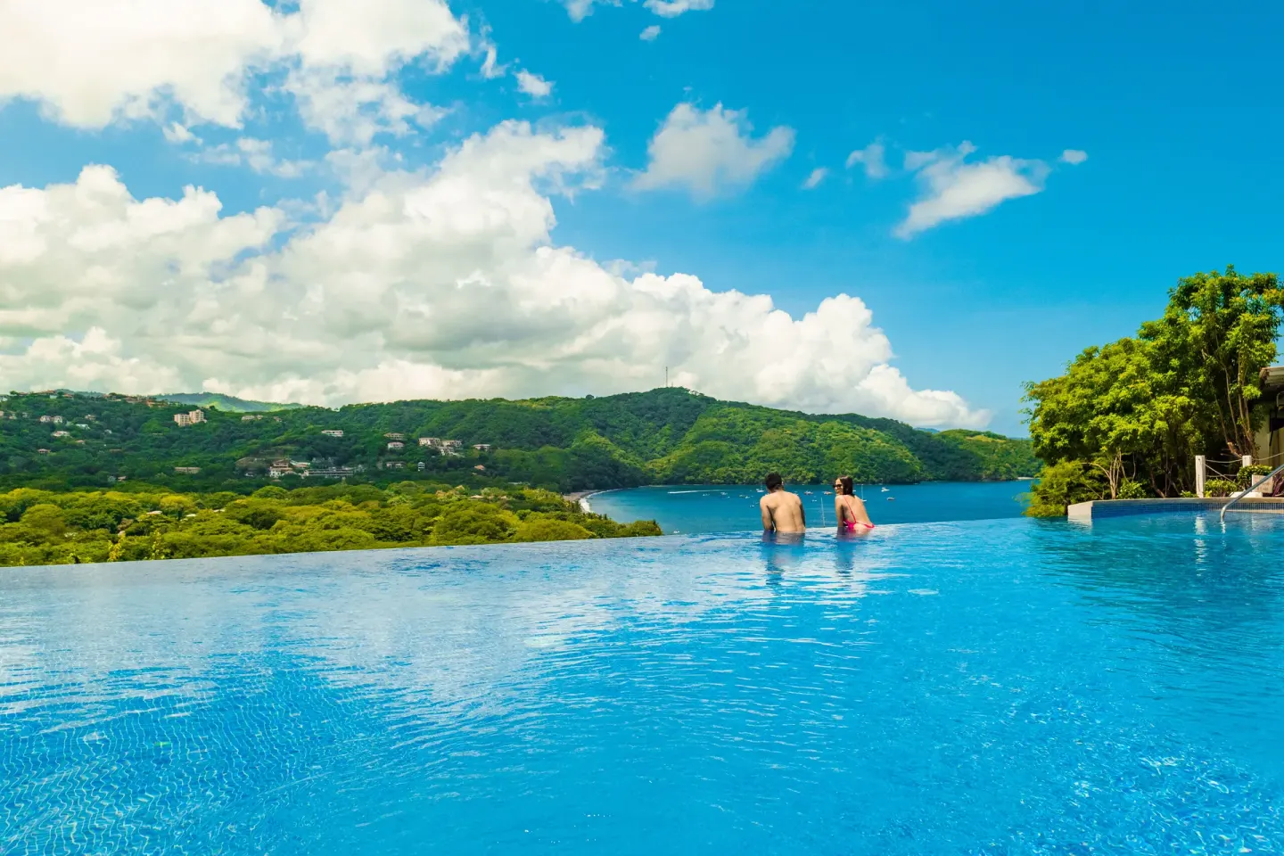 Wide infinity pool with two guests and green hillside view at Condovac la Costa.