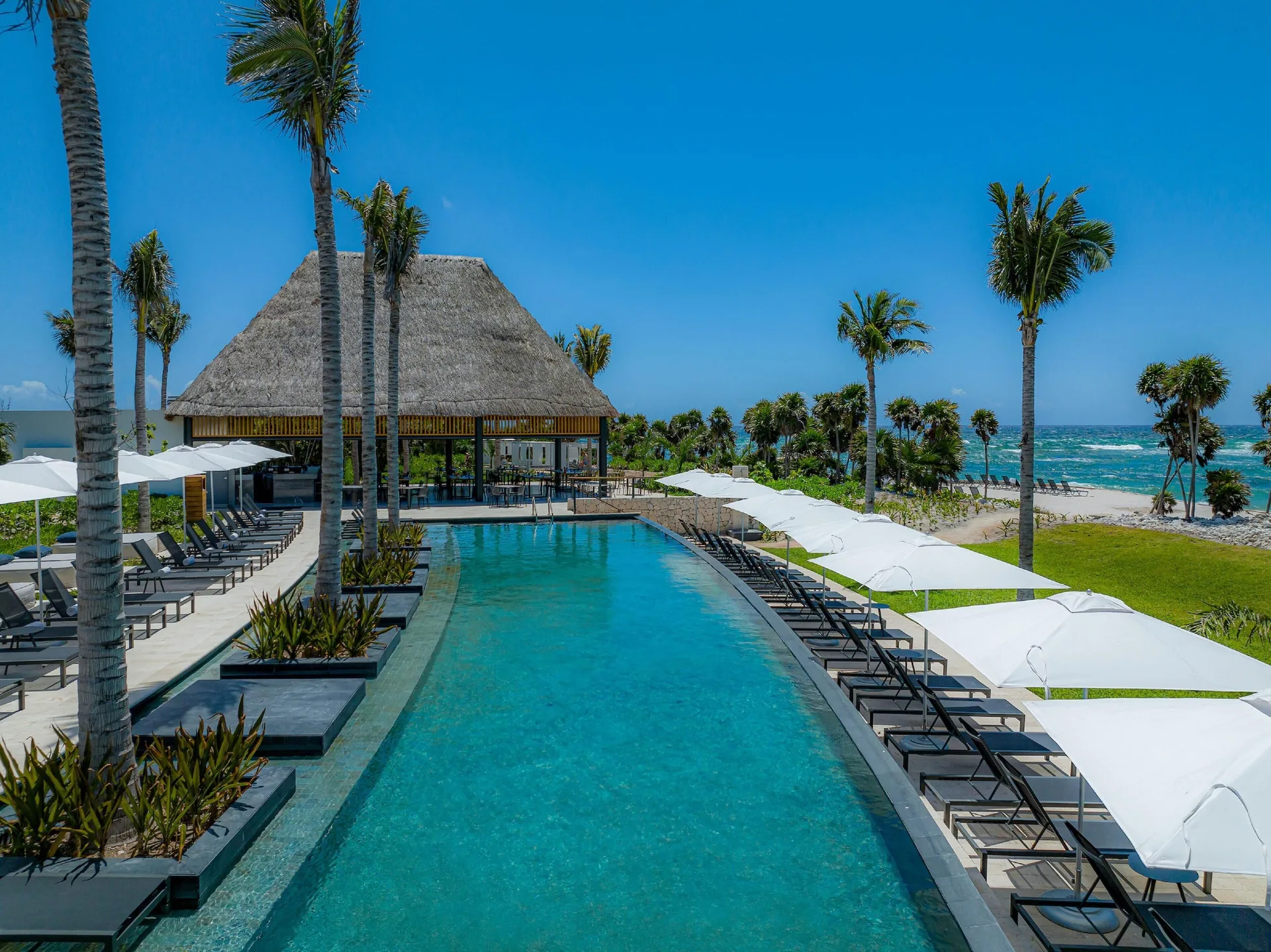 Long pool with lounge chairs, white umbrellas and ocean beyond at Conrad Tulum Riviera Maya.