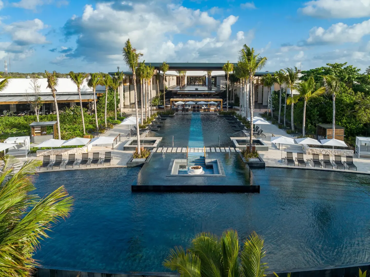 Resort pool at Conrad Tulum Riviera Maya with palm trees, loungers, umbrellas, and a central fire feature.