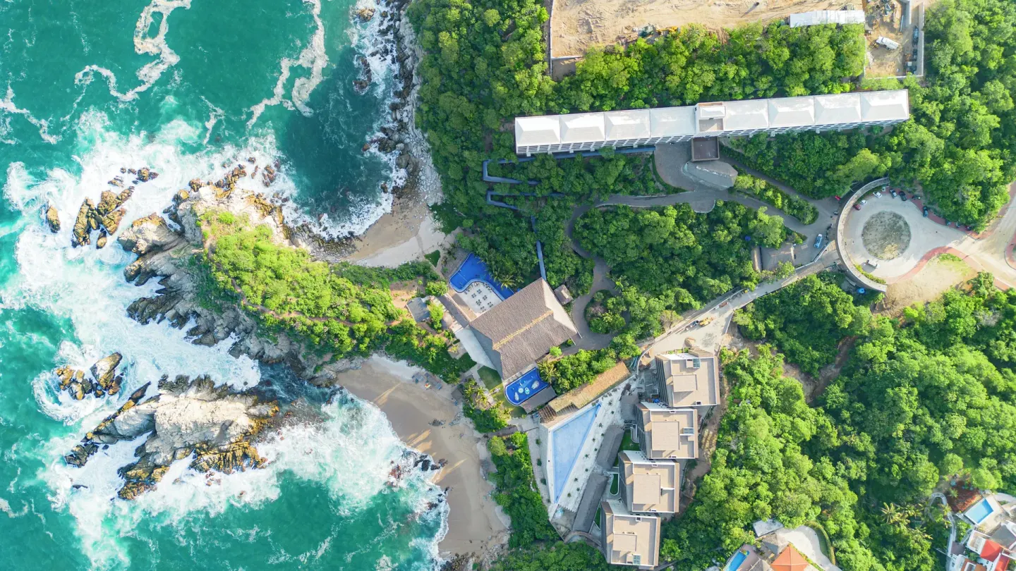 Aerial view of rocky coastline, beach coves, pools, and resort buildings at Coral Blue Huatulco.