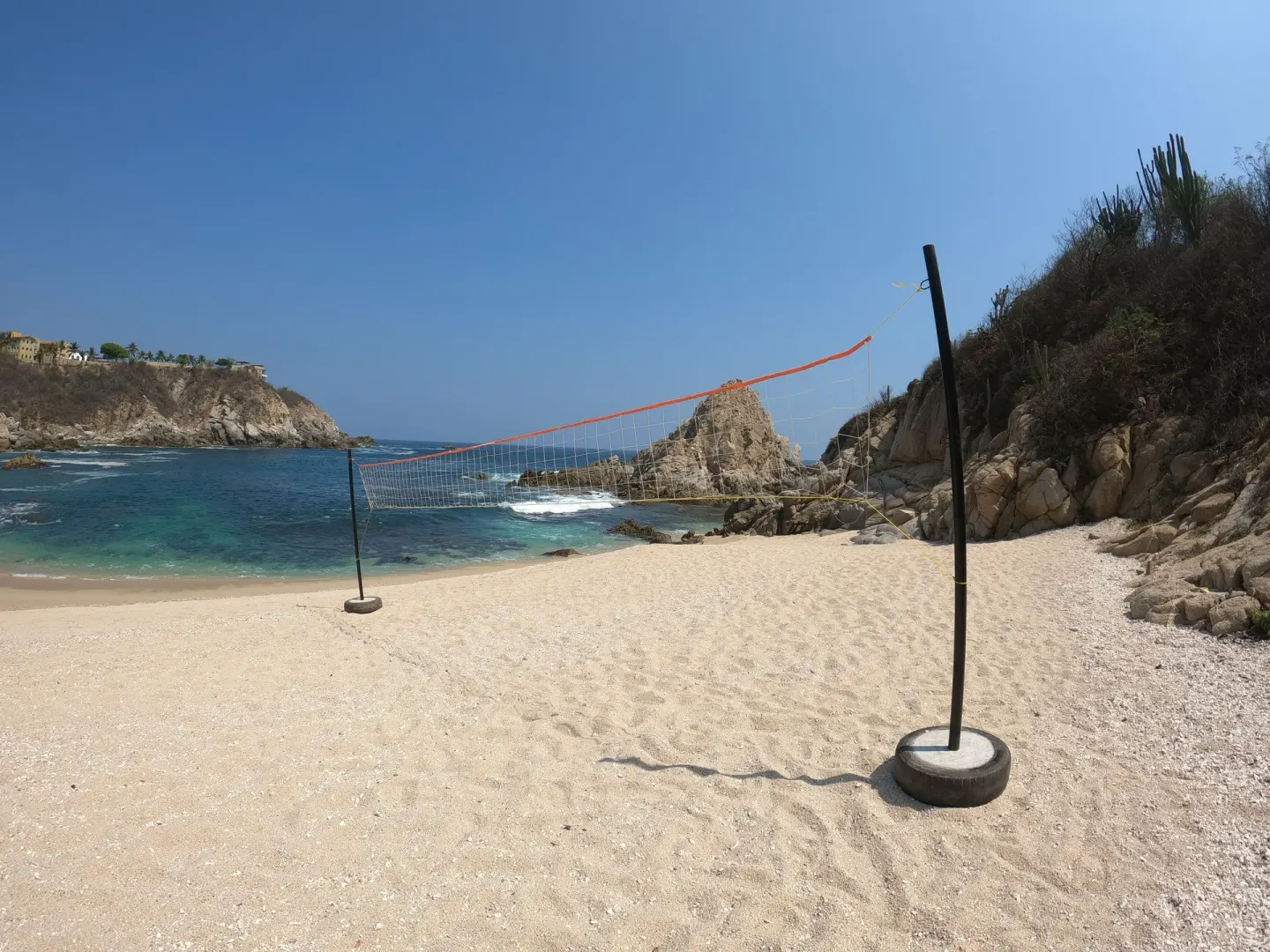 Beach volleyball net on a sandy cove beside rocky blue water at Coral Blue Huatulco.