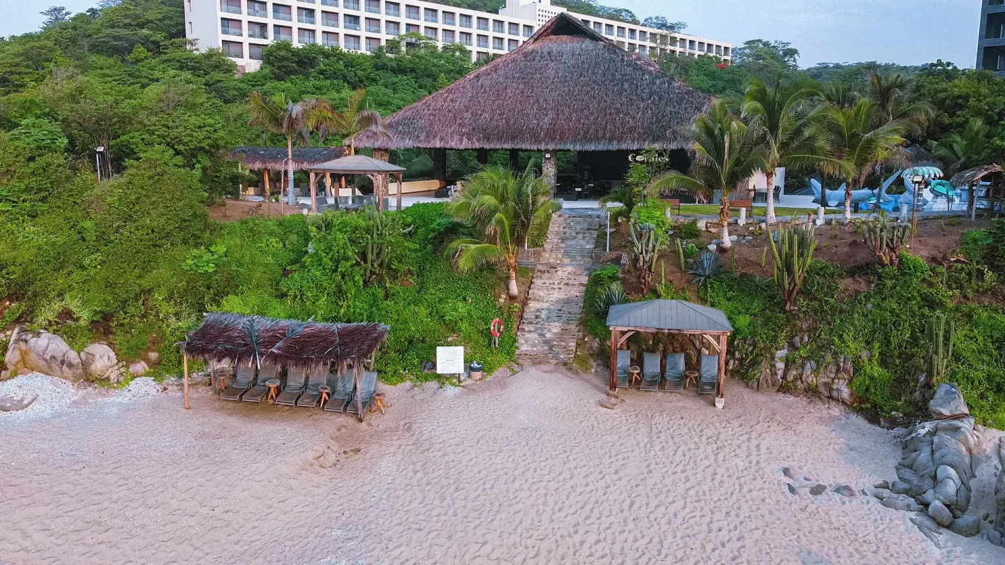Beachfront palapa, lounge shelters, palms, and resort building at Coral Blue Huatulco.