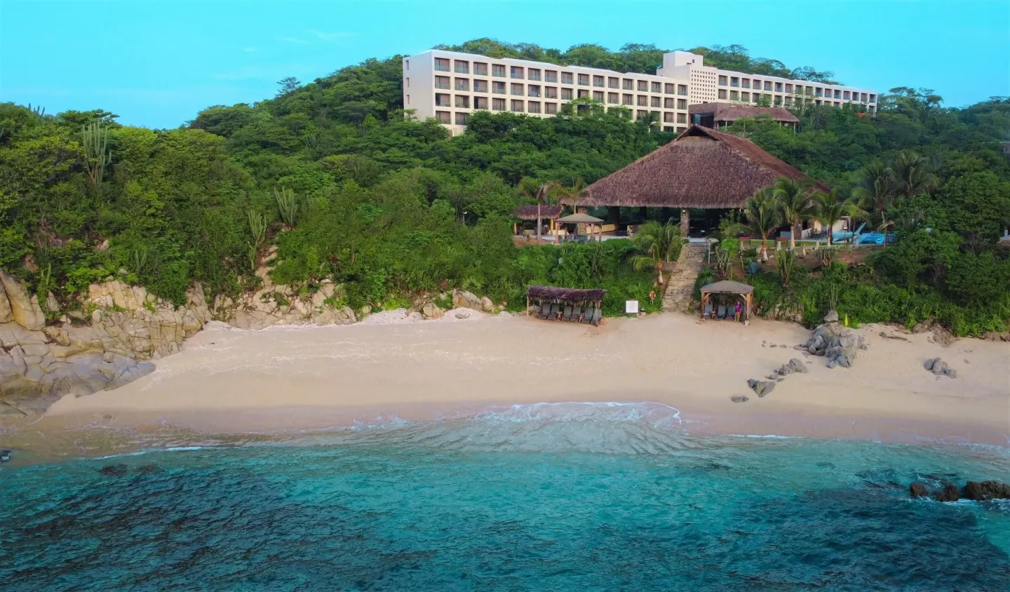 Beachfront view of Coral Blue Huatulco with turquoise water, sand, palms, and hillside rooms.