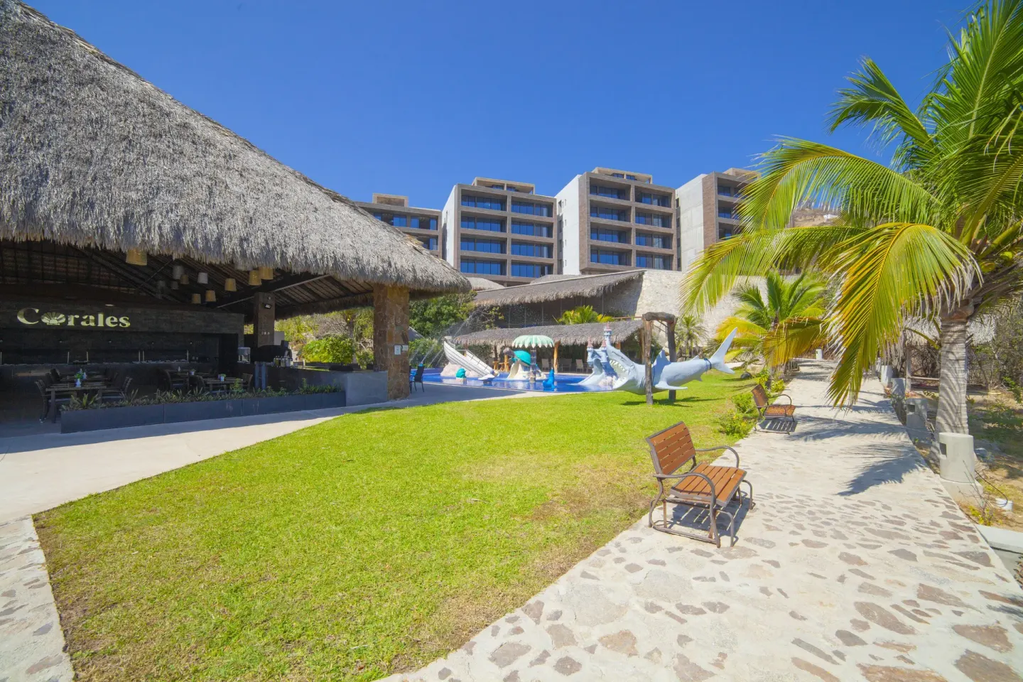 Garden lawn, thatched bar, pool area, and hotel buildings at Coral Blue Huatulco.