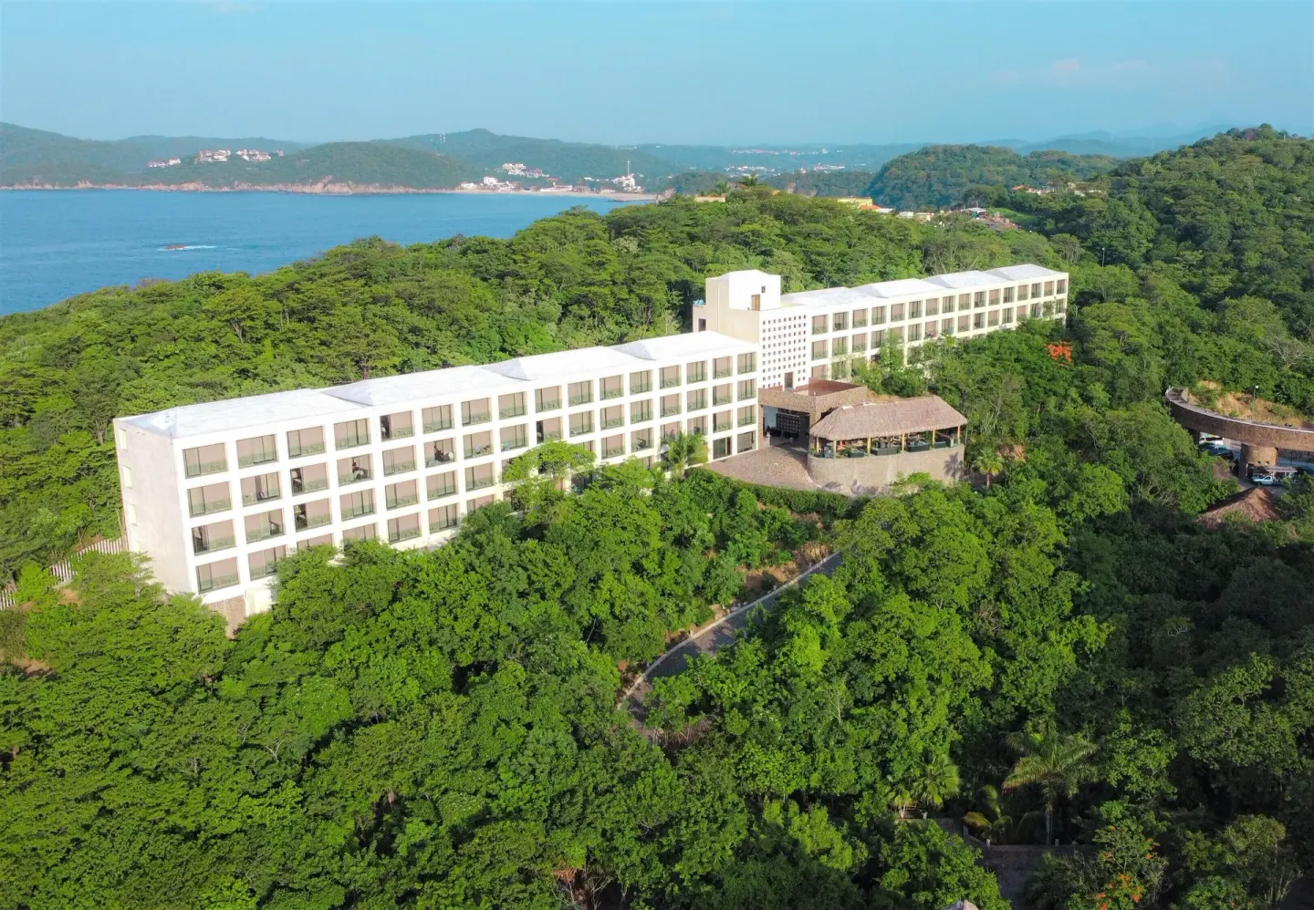 Aerial view of Coral Blue Huatulco building surrounded by forested hills and bay coastline.