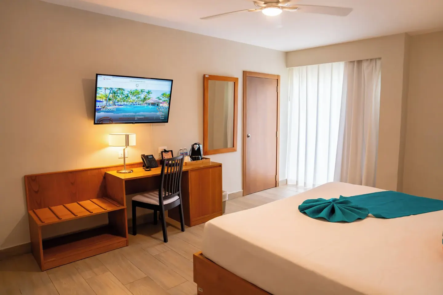 Deluxe Room bed with desk, mirror, TV, and curtains at Coral Costa Caribe Beach Resort.