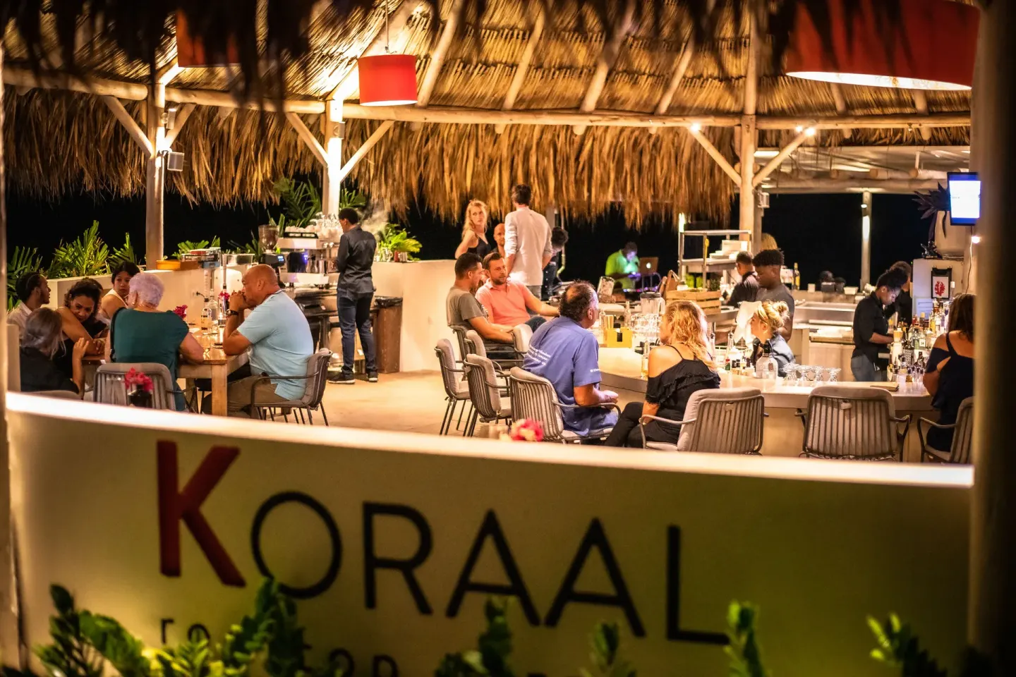 Koraal Curaçao evening bar with seated guests under a thatched roof at Coral Estate Luxury Resort.