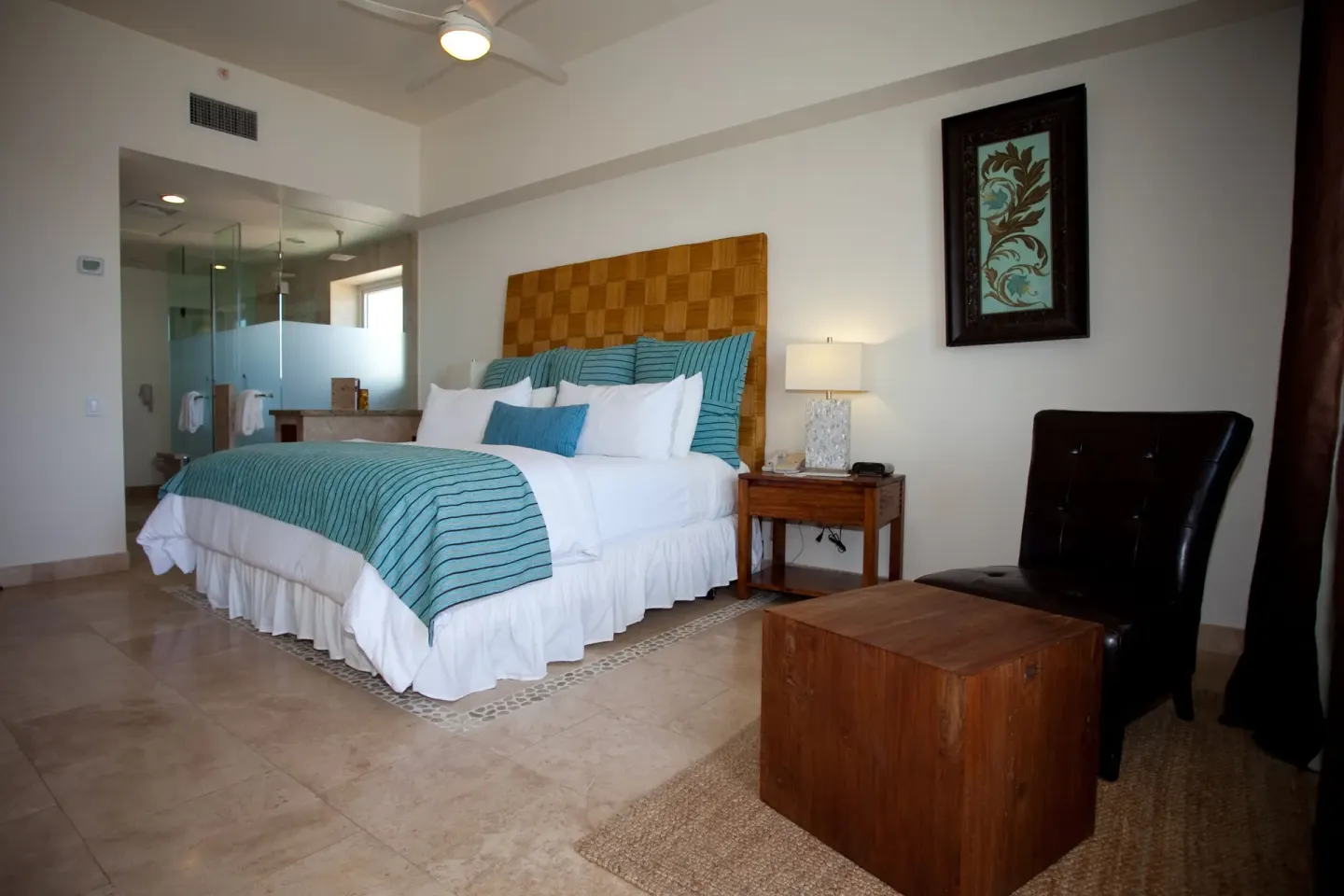 King bed guest room with sitting chair, wood ottoman, and glass bathroom at Corazón Cabo.