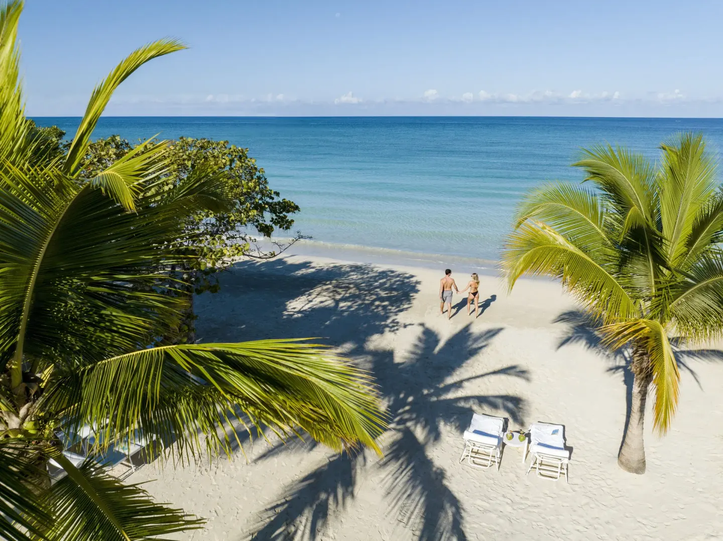 Palm-framed beach with two loungers and a couple walking at Couples Negril.