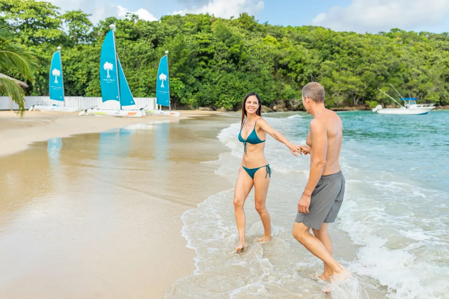 A couple walks in the surf with small sailboats on the beach at Couples Sans Souci.