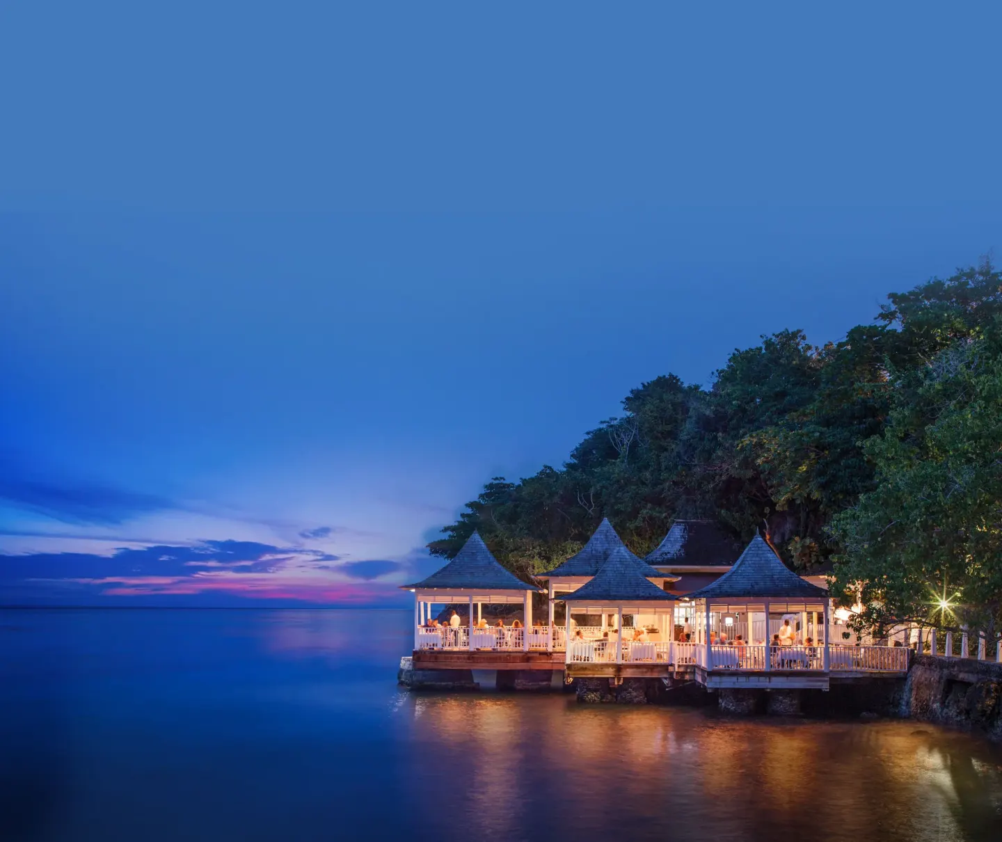 An illuminated overwater restaurant sits beside the sea at dusk at Couples Tower Isle.