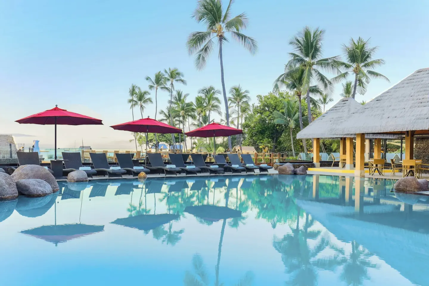 Pool at Courtyard King Kamehameha's Kona Beach Hotel with lounge chairs, red umbrellas, and palms.