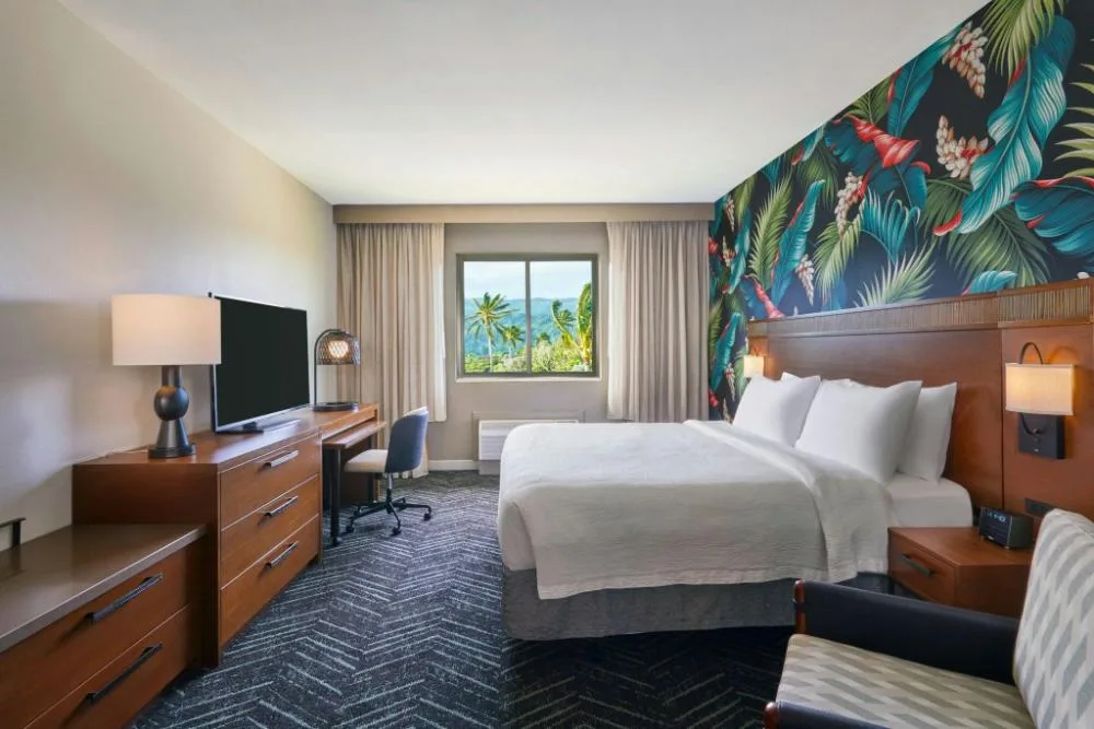 Bedroom with king bed, tropical accent wall, desk, TV, and window view at Courtyard Oahu North Shore.