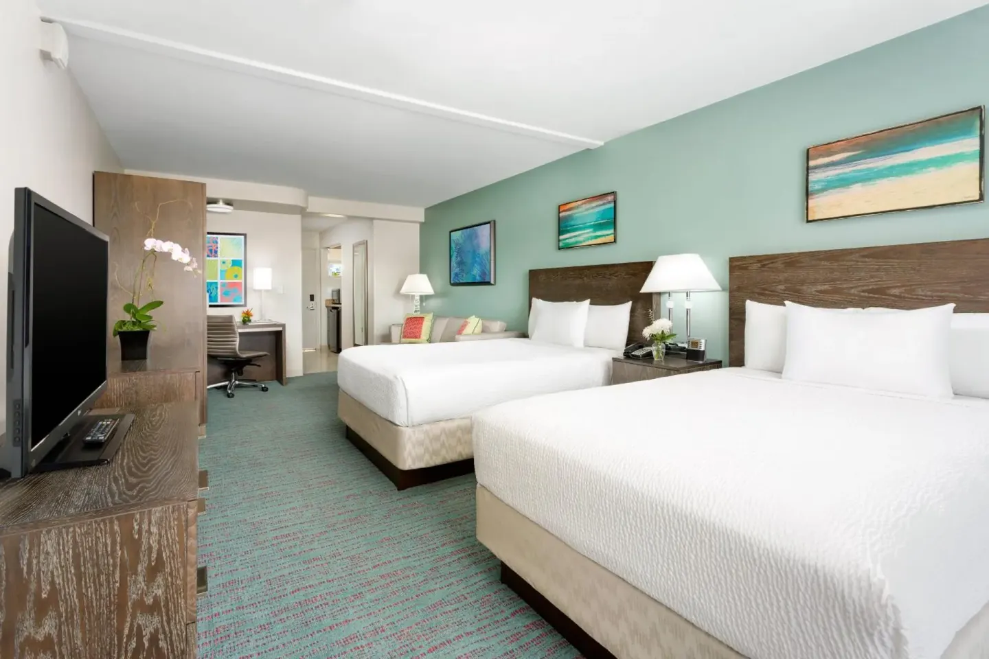 Guest room with two beds, TV, desk, sofa, and beach art at Courtyard Waikiki Beach.