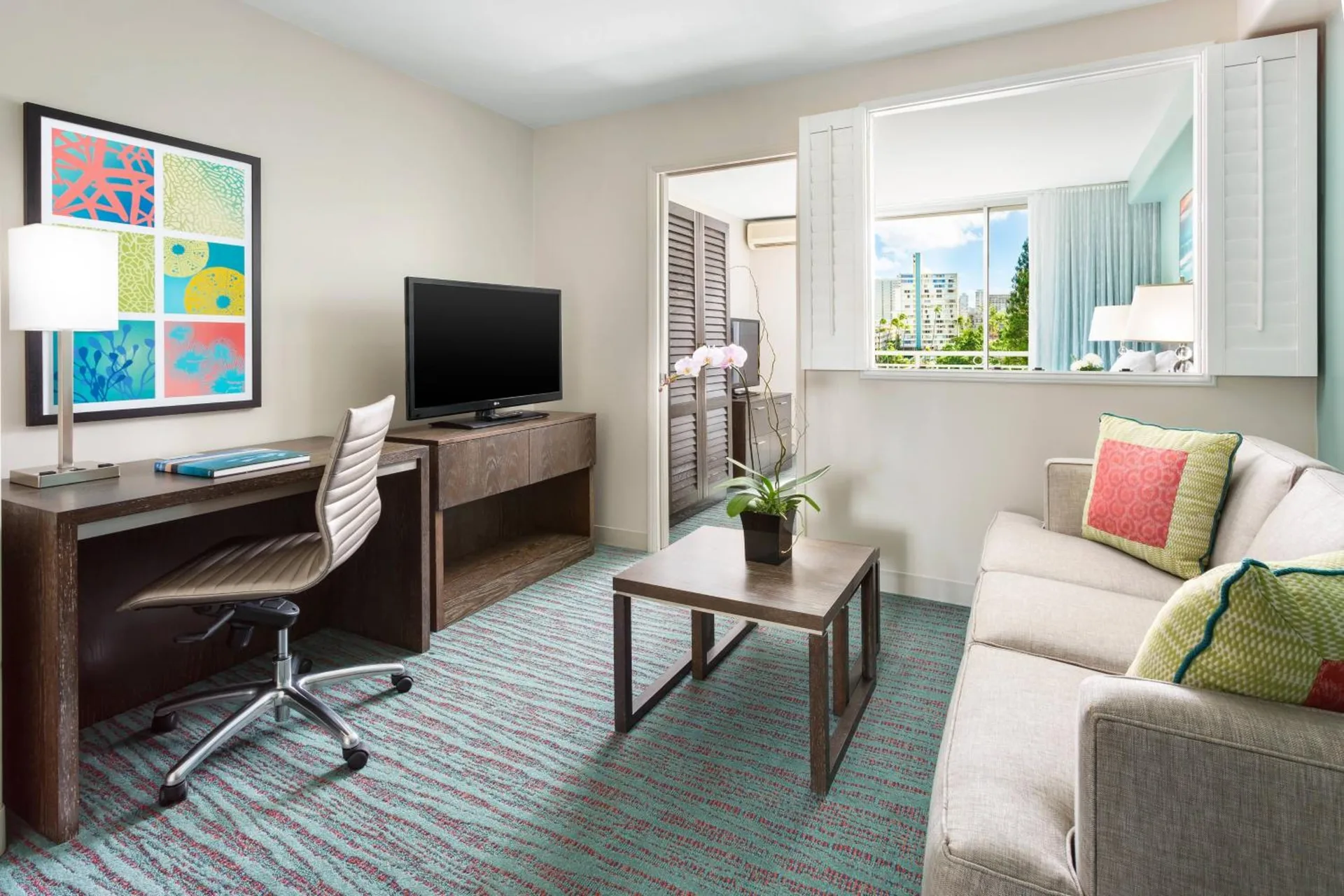 Suite living area with sofa, desk, TV, and interior window at Courtyard Waikiki Beach.