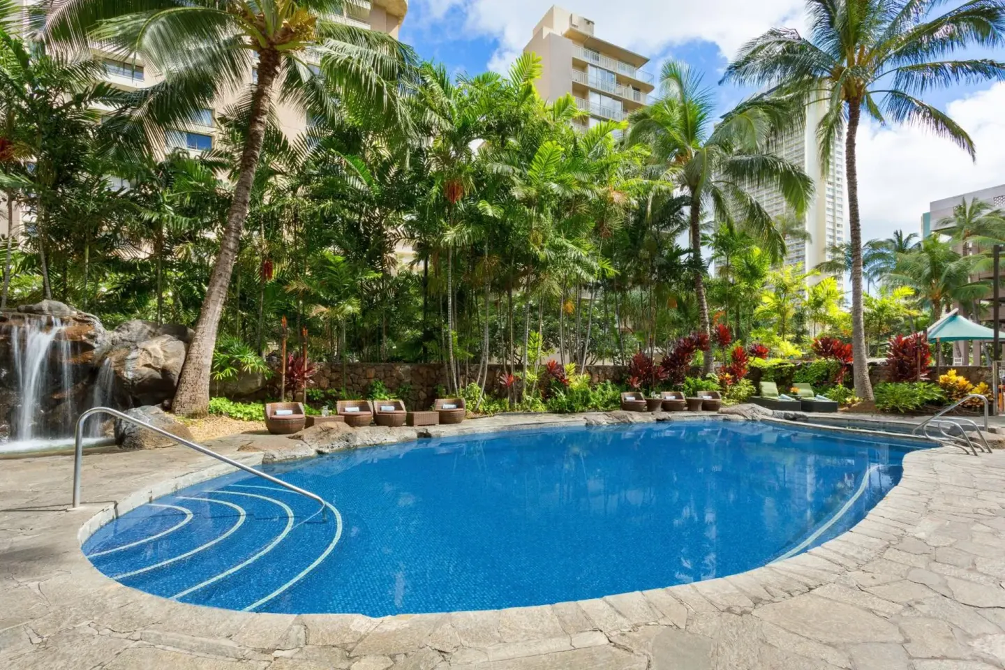 Outdoor pool with palm trees, lounge chairs, and a waterfall at Courtyard Waikiki Beach.