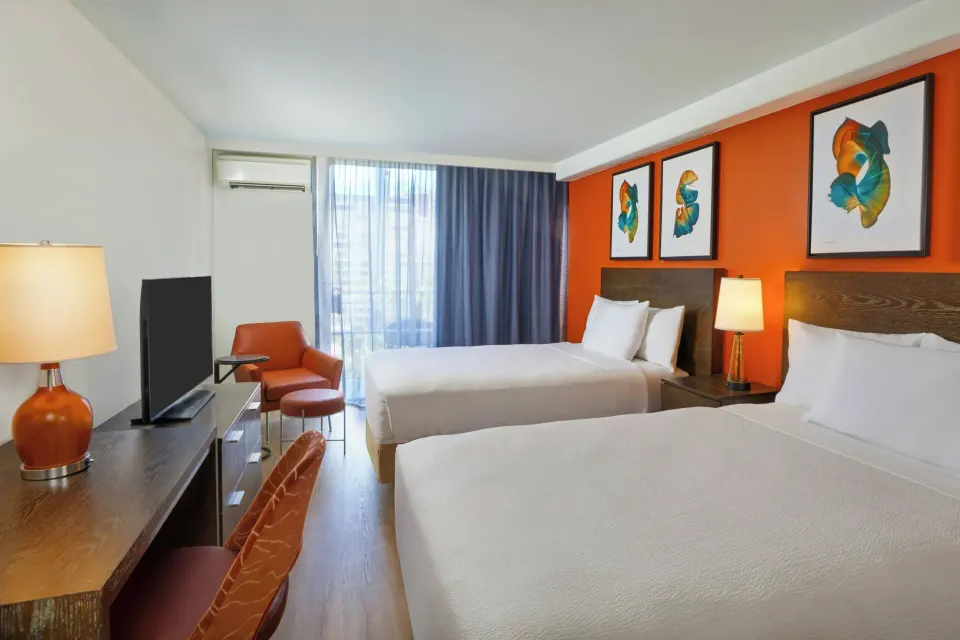 Deluxe balcony room with two beds, orange wall art, desk, TV, and seating at Courtyard Waikiki Beach.