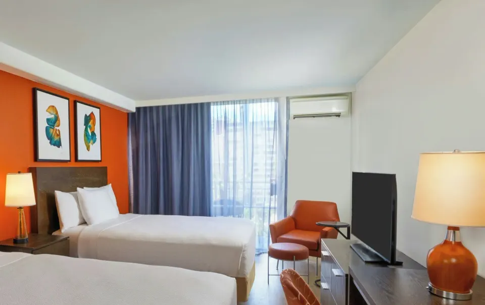 Deluxe balcony room with two beds, armchair, TV, lamps, and draped window at Courtyard Waikiki Beach.
