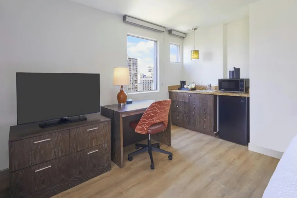 Deluxe balcony room work area with TV, desk, kitchenette, and window at Courtyard Waikiki Beach.