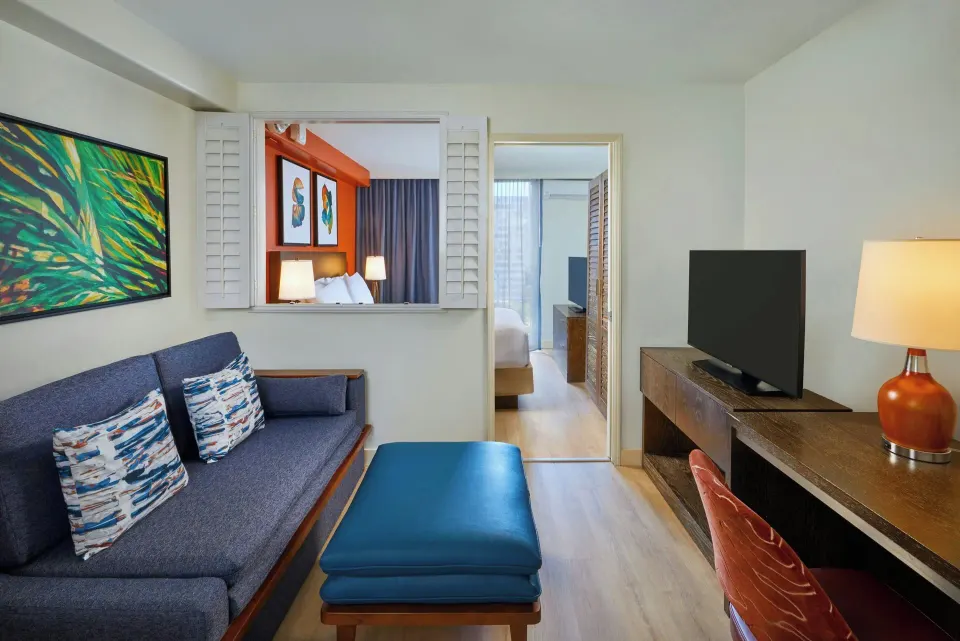 Suite living room with sofa, ottoman, desk, TV, and bedroom beyond at Courtyard Waikiki Beach.