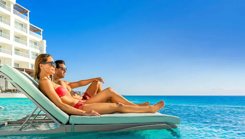Couple relaxing on in-pool loungers with hotel building and sea at Cozumel Palace