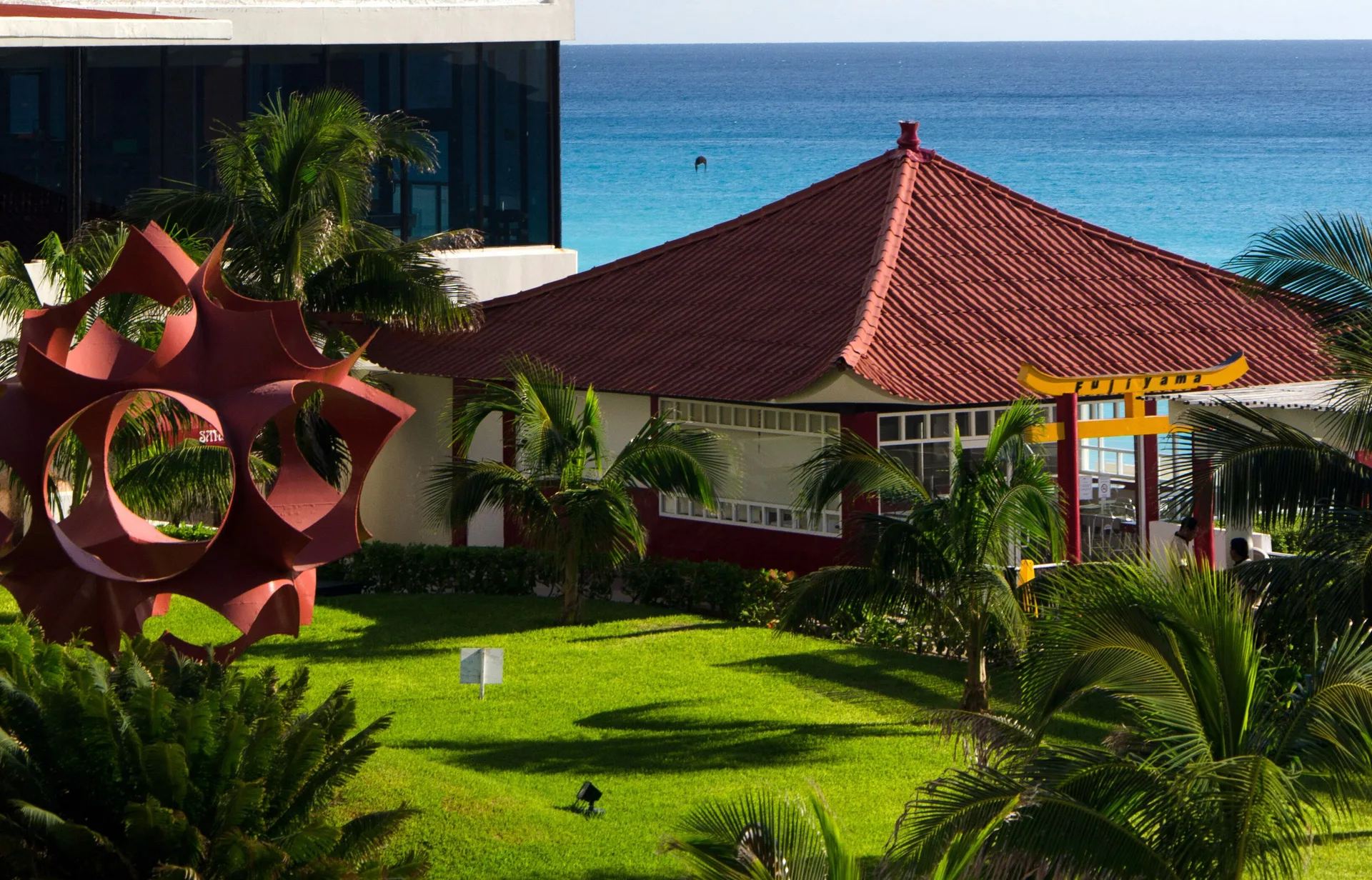 Oceanfront lawn with palms, sculpture, and red-roofed building at Crown Paradise Club Cancun.